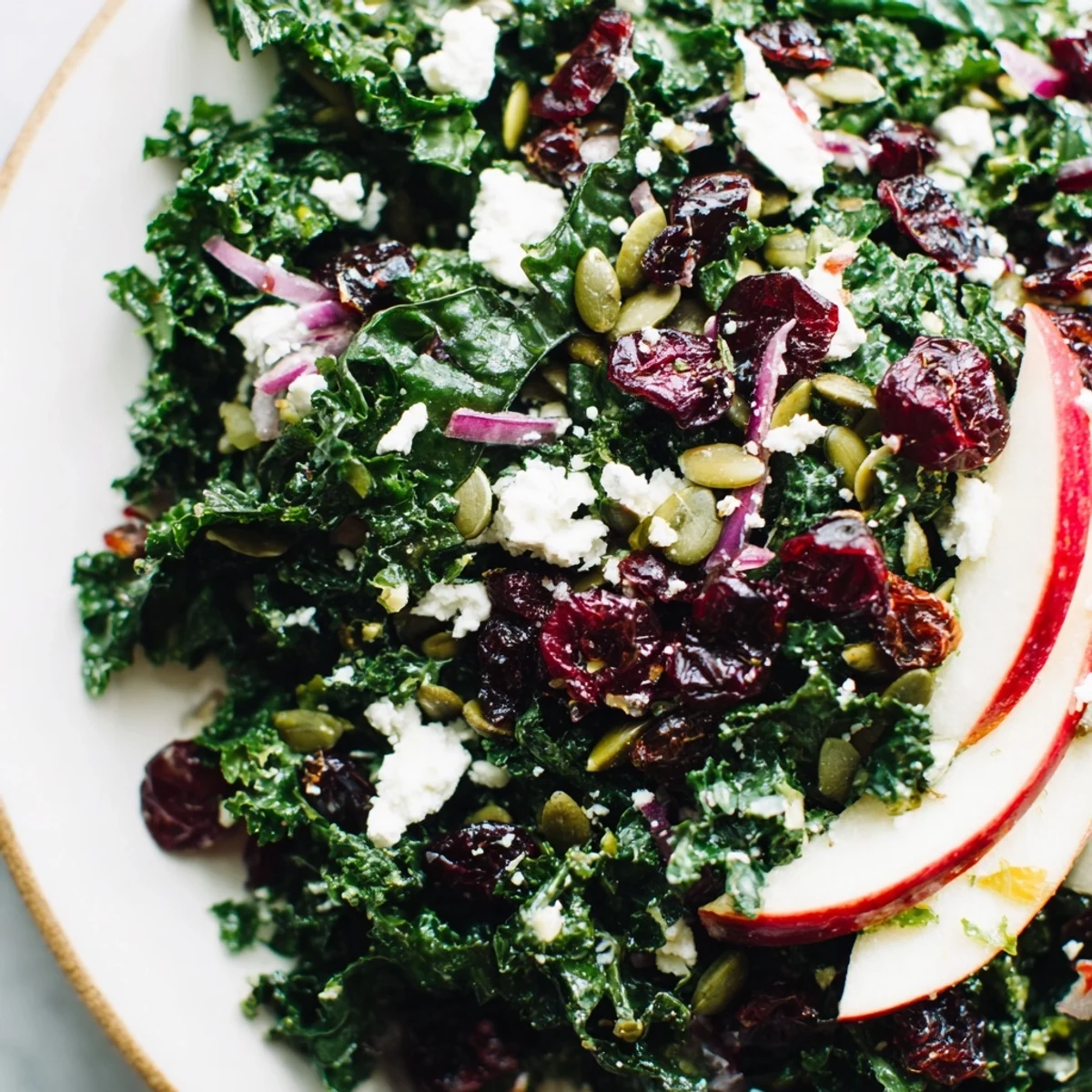 A close-up of Winter Kale Salad with cranberries and seeds, drizzled with lemony dressing, topped with apple slices and feta.