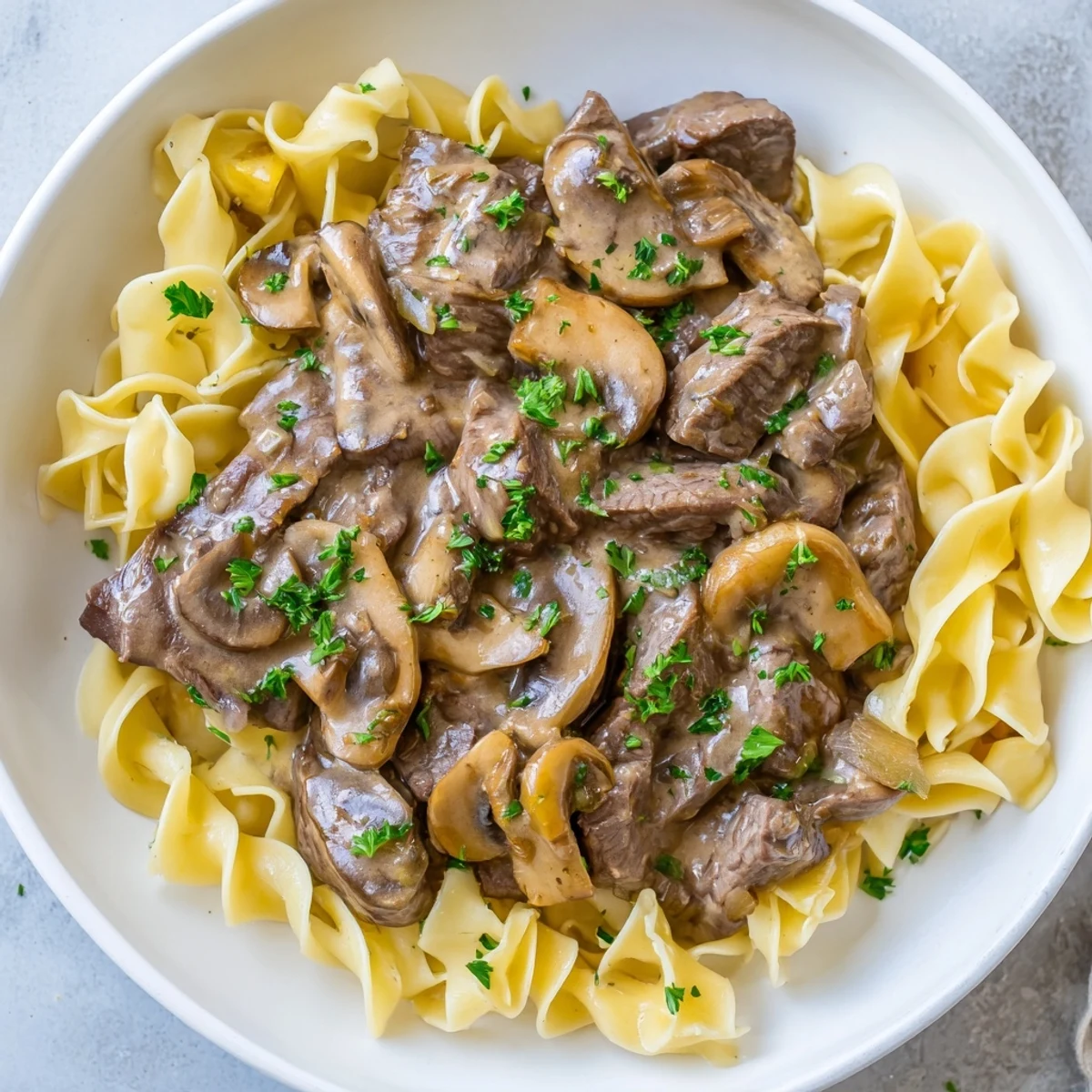 Beef stroganoff with egg noodles served in a white bowl garnished with fresh parsley.