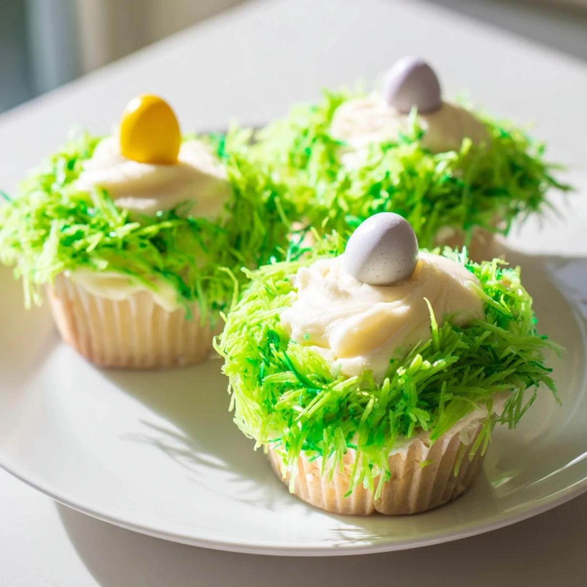 A close-up of Easter Basket Cupcakes with Coconut Grass displays pastel frosting, shredded green coconut, and tiny chocolate eggs for a sweet holiday treat.