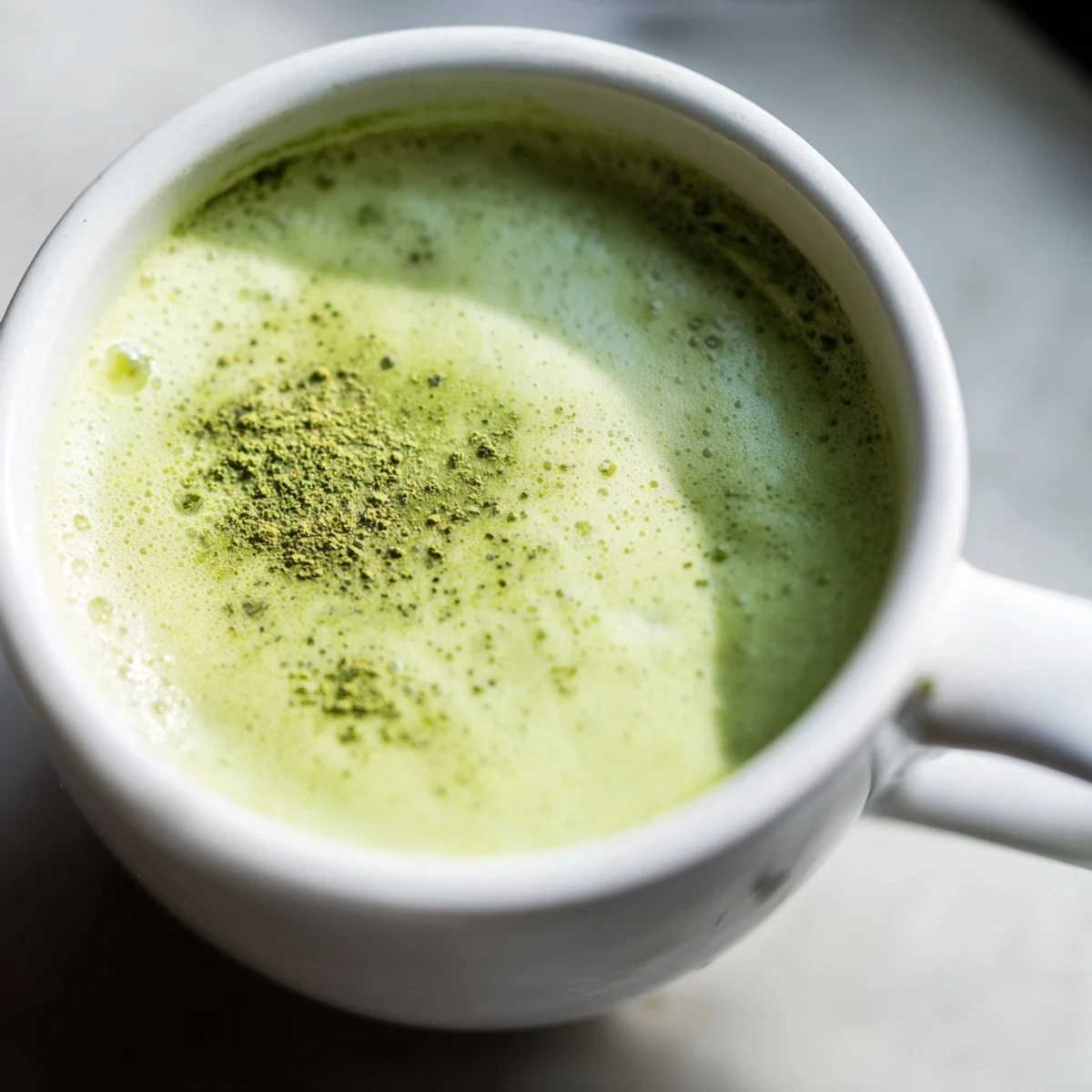 Vibrant green matcha latte in a white mug topped with a creamy almond milk foam and a light dusting of matcha powder.